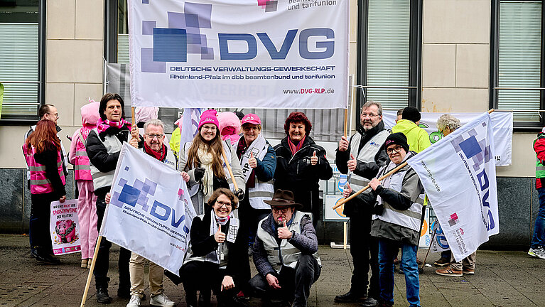 Das Bild zeigt eine Gruppe von Menschen, die an einem Protest teilnehmen. Im Mittelpunkt stehen mehrere Personen, die Banner und Fahnen mit dem Logo der Deutschen Verwaltungs-Gewerkschaft (DVG) tragen. Auf dem großen Banner im Hintergrund steht deutlich "DVG" und darunter der vollständige Name der Organisation sowie ihre Zugehörigkeit zum dbb beamtenbund und tarifunion. Die Website der DVG-RLP ist ebenfalls angegeben.