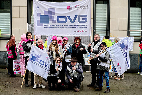 Das Bild zeigt eine Gruppe von Menschen, die an einem Protest teilnehmen. Im Mittelpunkt stehen mehrere Personen, die Banner und Fahnen mit dem Logo der Deutschen Verwaltungs-Gewerkschaft (DVG) tragen. Auf dem großen Banner im Hintergrund steht deutlich "DVG" und darunter der vollständige Name der Organisation sowie ihre Zugehörigkeit zum dbb beamtenbund und tarifunion. Die Website der DVG-RLP ist ebenfalls angegeben.